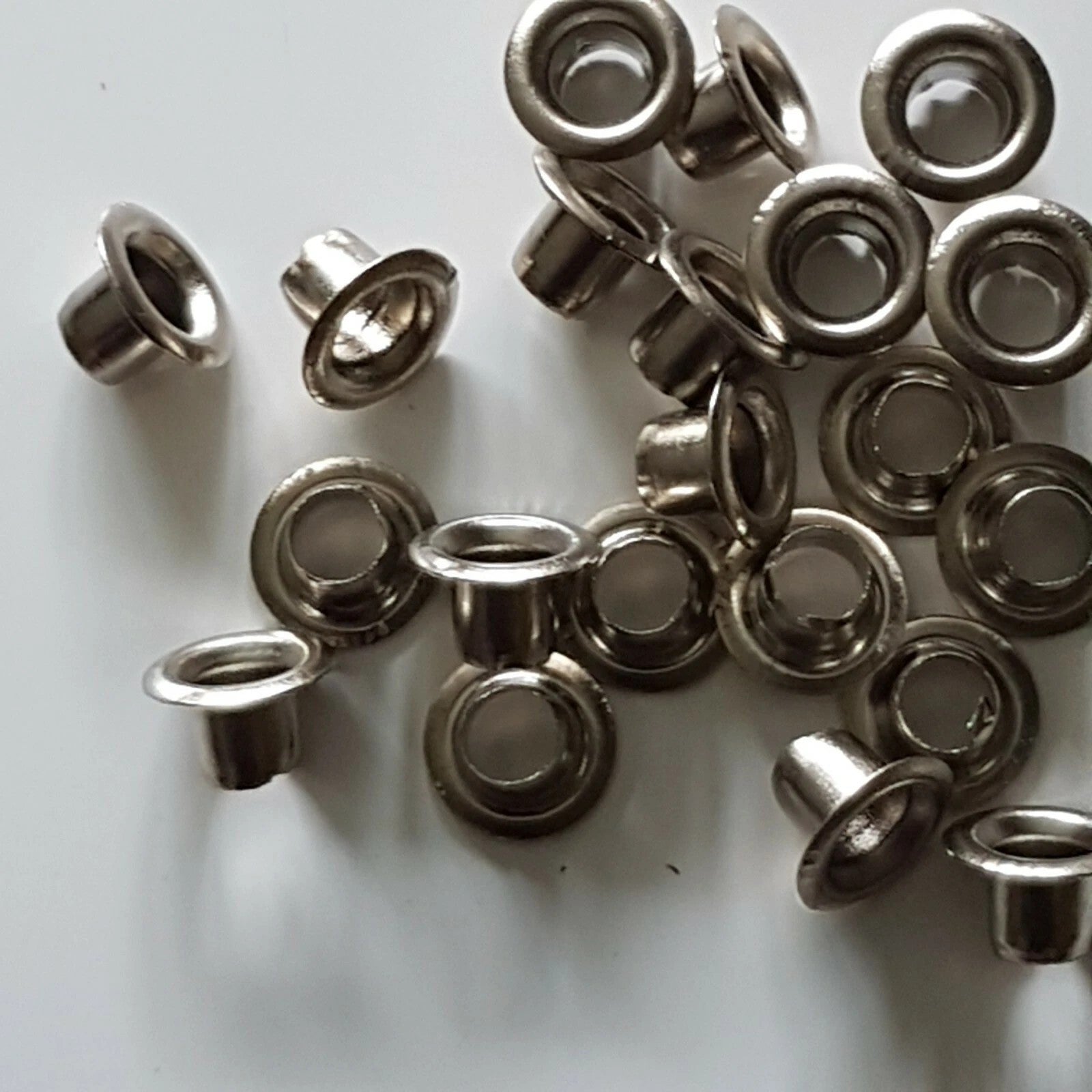 A pile of small metallic eyelets with a silver finish, scattered on a plain white surface.