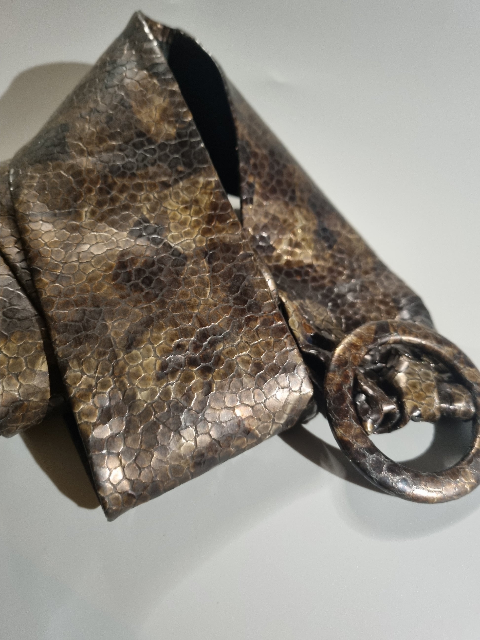 A close-up of a textured metallic belt with a hammered, scale-like finish in bronze and brown tones, featuring a large circular ring buckle.