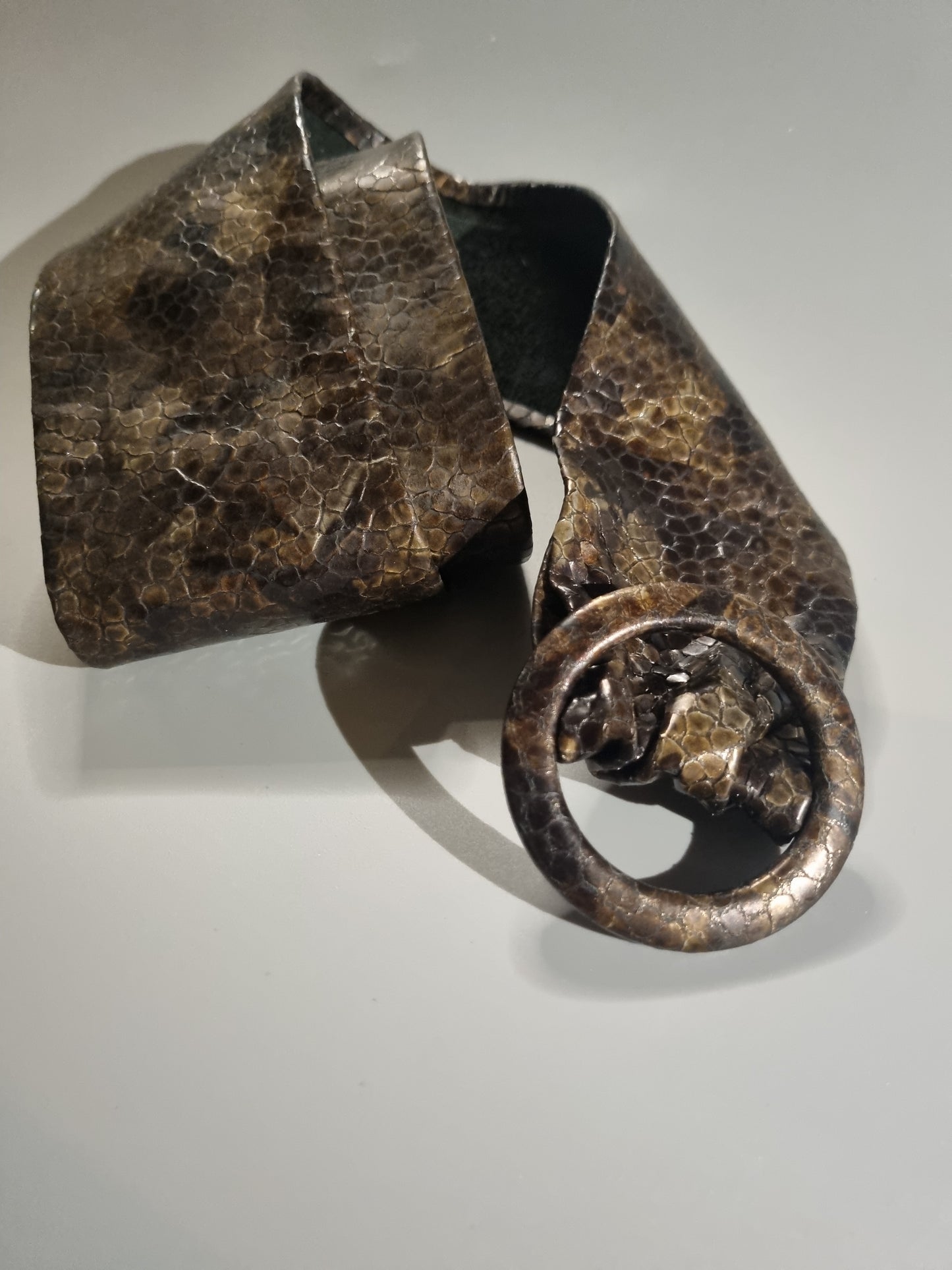 A sculptural hammered metal belt with a textured, oxidized finish in dark bronze and brown tones, featuring a wide strap and a circular buckle.