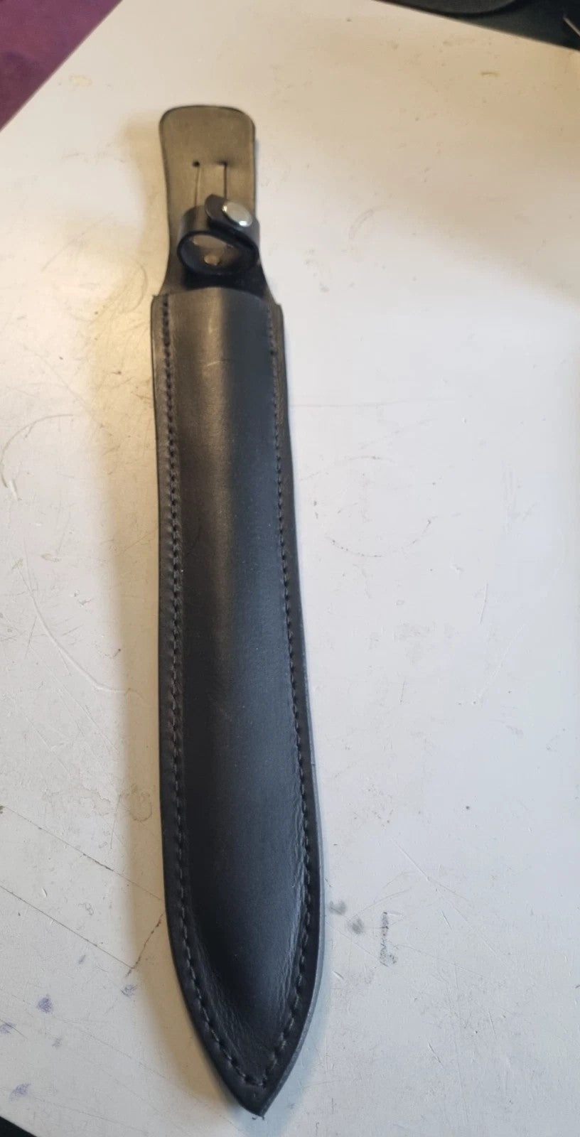 A black leather knife sheath with a handle snap loop p, shown on a light-colouredsurface