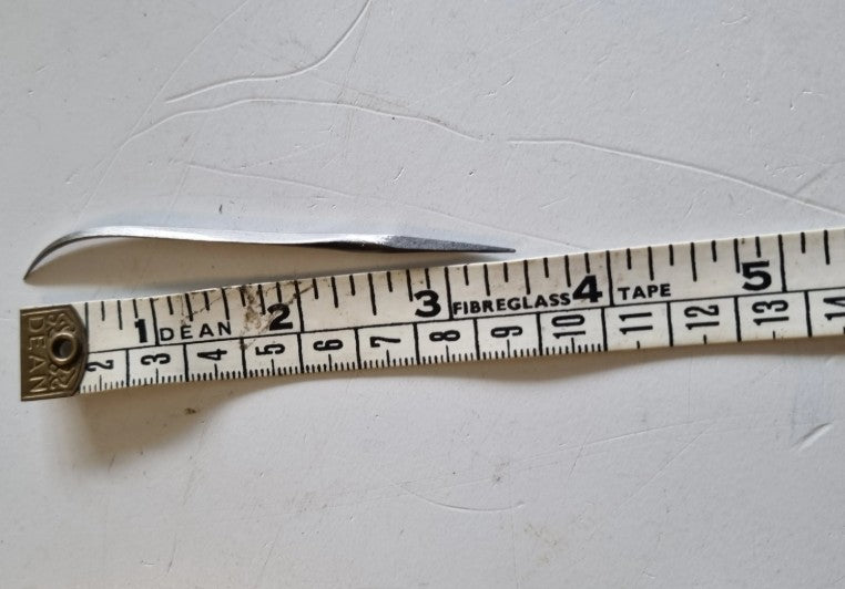 A curved diamond awl tool placed above a measuring tape on a light off-white surface. The tape shows markings including 'FIBREGLASS TAPE' and numbers up to 3.5 inches.