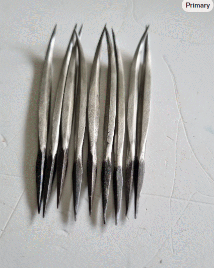 A group of long, slender cast metal diamond awls with a pointed tip lies vertically on a gray wood-grain surface.
