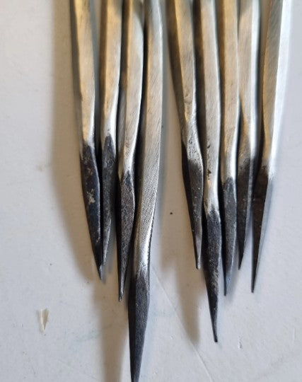 A close-up of several cast steel metal diamond awls ends showing forged ends arranged vertically on a light off-white surface.