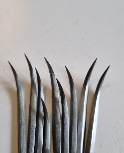 A close-up of several long, curved diamond awls with pointed tips arranged vertically on a plain off-white surface.