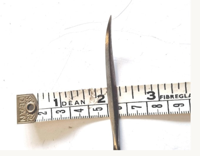 A close-up of a silver cast metal hand sewing diamond curved awl lying horizontally over a white measuring tape, showing its width size relative to the tape measure.