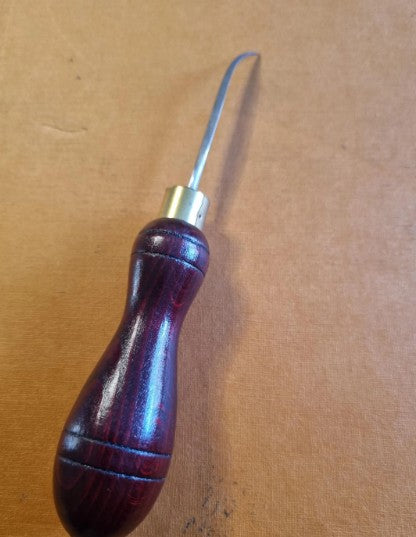 A leatherworking tool with a dark reddish-brown wooden handle and a curved silver cast steel diamond curved point, resting on a brown textured surface.