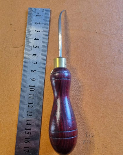 A leatherworking tool with a dark reddish-brown wooden handle and a curved diamond silver metal point awl, shown next to a metal ruler on a brown leather background.