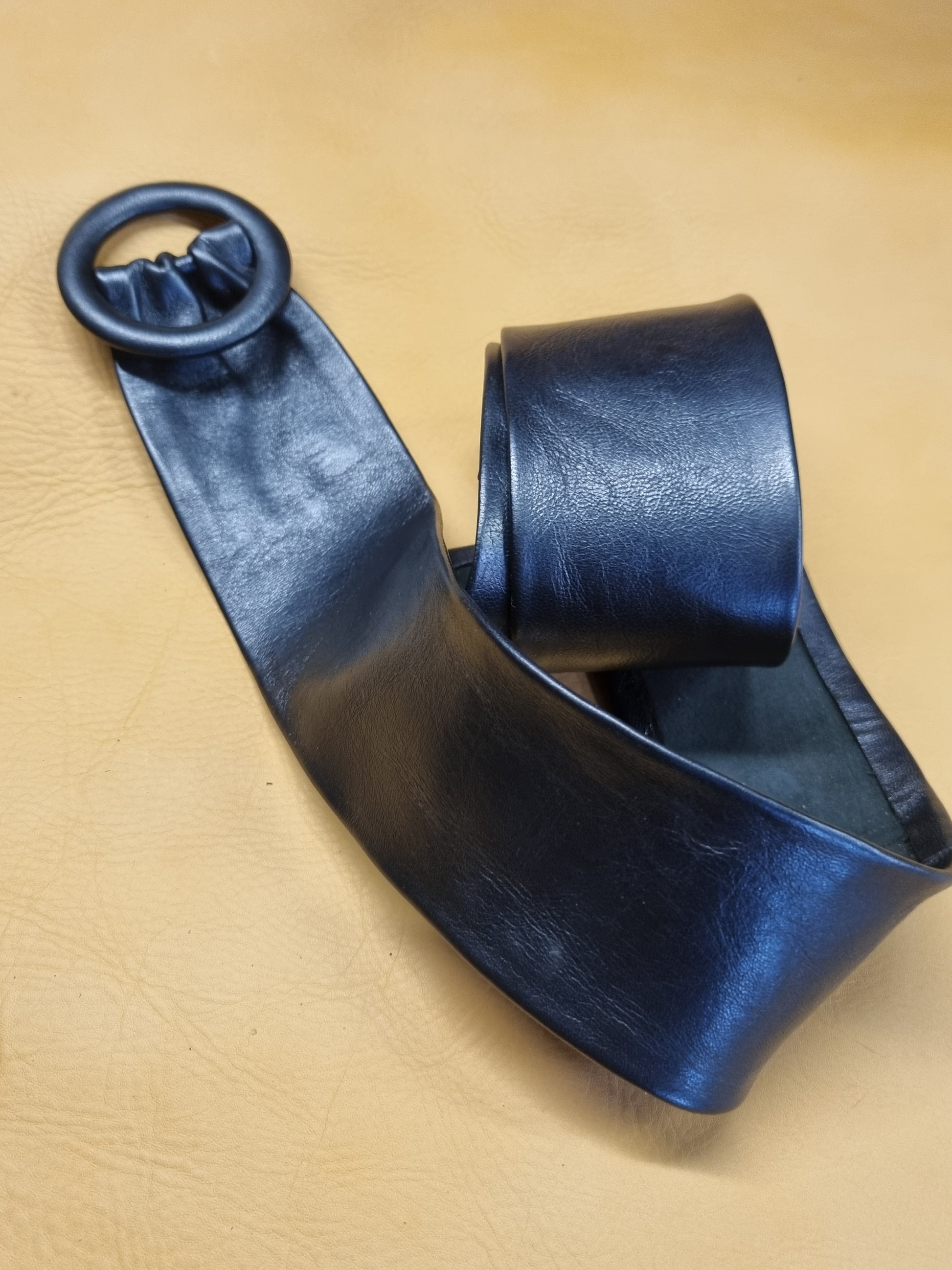 A navy blue cinch leather belt with a wide strap and a circular leather covered buckle, displayed on a textured tan leather background.