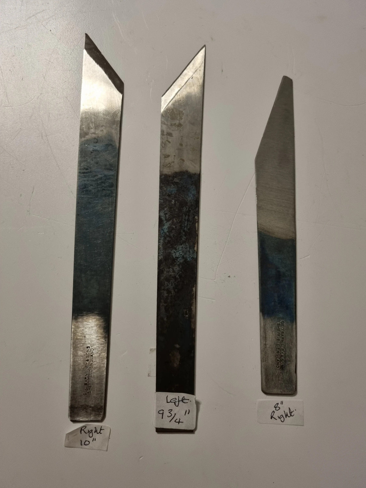 Three long, narrow metal knife blades or cutting tools displayed vertically on a light gray surface, with handwritten size labels below each piece.