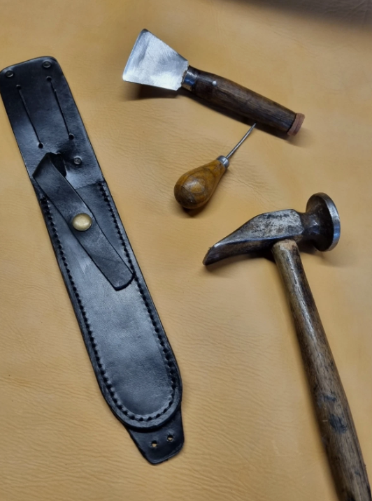 A leather dagger sheath with a snap closure lies on a tan leather surface alongside a hammer, a small chisel, and a small hand tool with a wooden handle.
