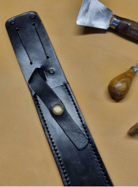A black leather knife sheath with a brass snap button, shown on a tan leather background with hand tools partially visible.