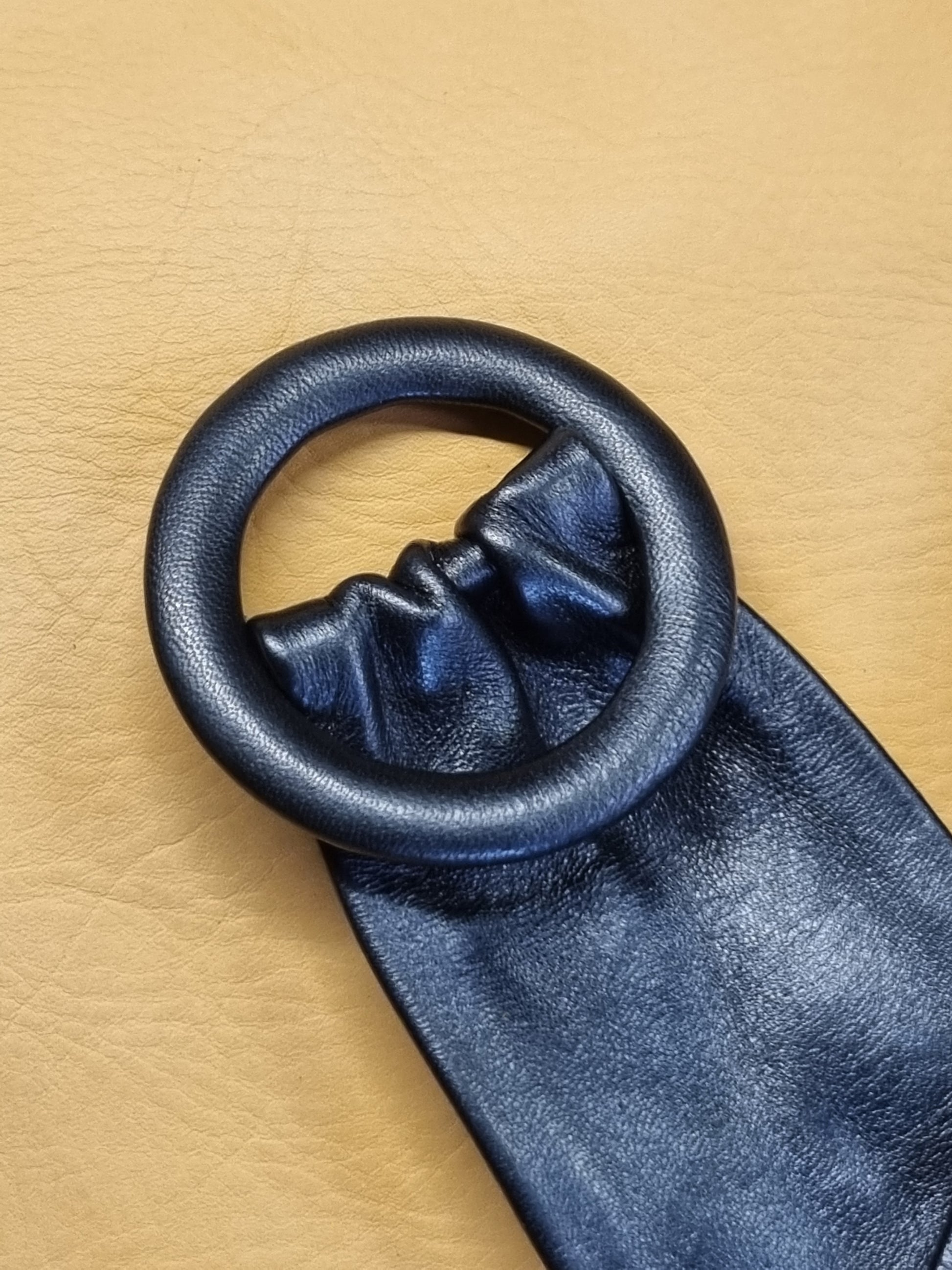 A close-up of a dark navy blue leather belt with a large, circular, matte leather covered buckle, laid diagonally on a textured tan leather background.