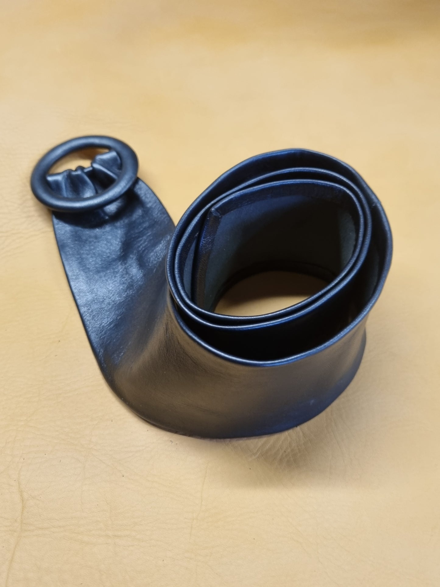 Close of the navy blue leather belt with a wide strap and a large circular leather covered buckle, coiled on a tan textured leather background.