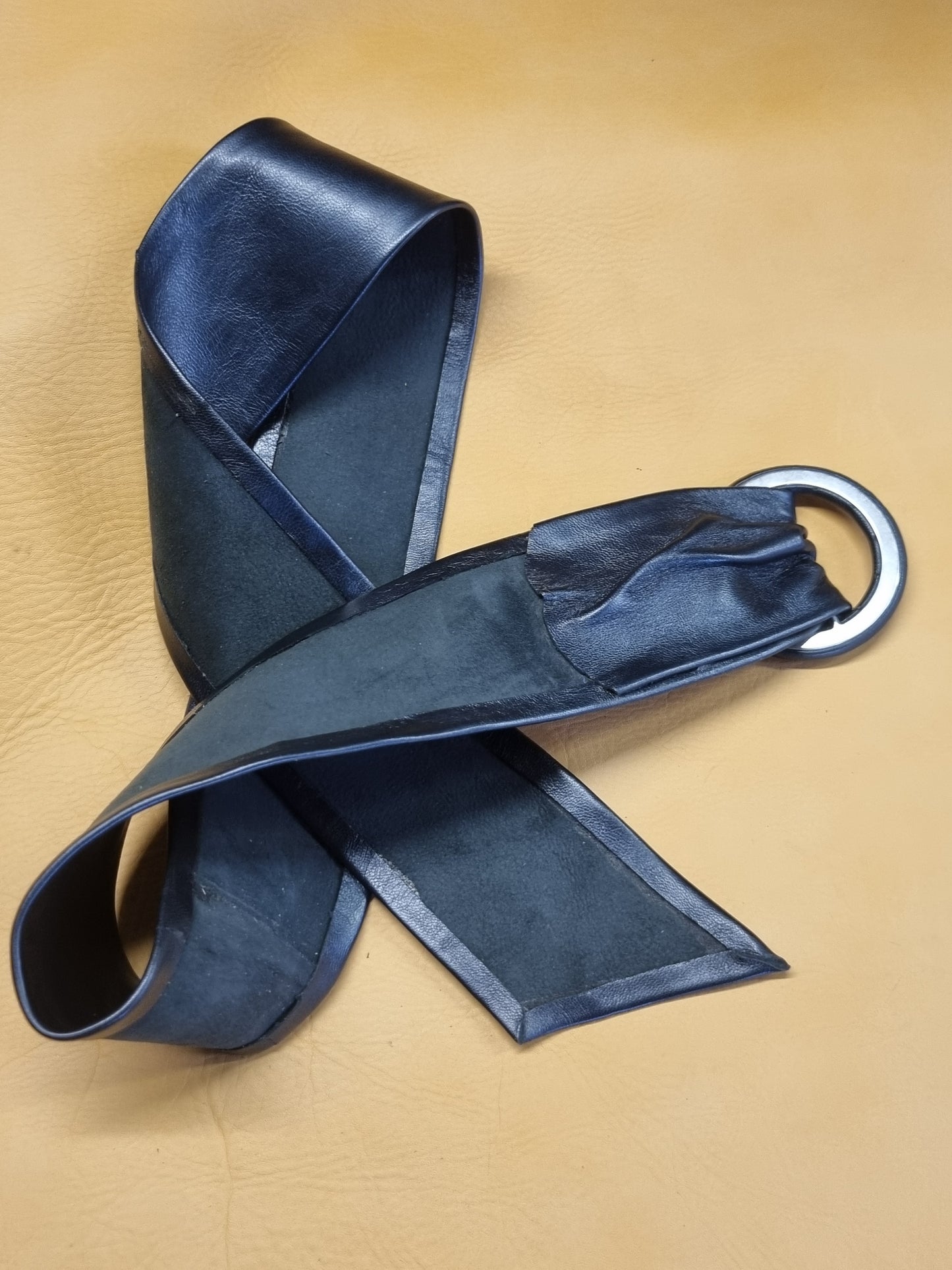 A dark navy blue leather belt turned over to reveal finish, featuring a silver metal D-ring, arranged on a textured tan leather background.