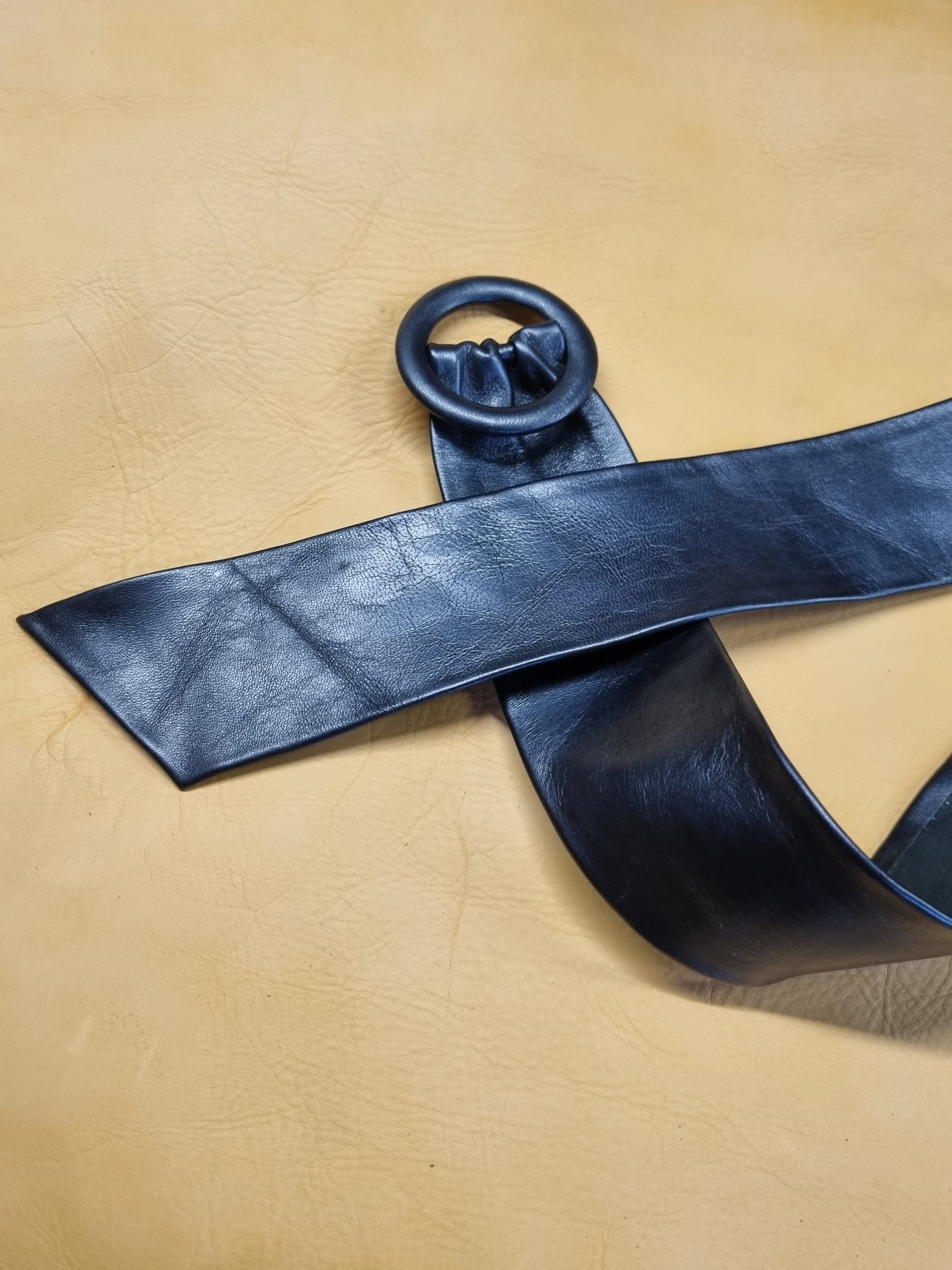 A wide navy blue leather belt with a large circular leather covered buckle shown laid diagonally across a textured tan leather background.