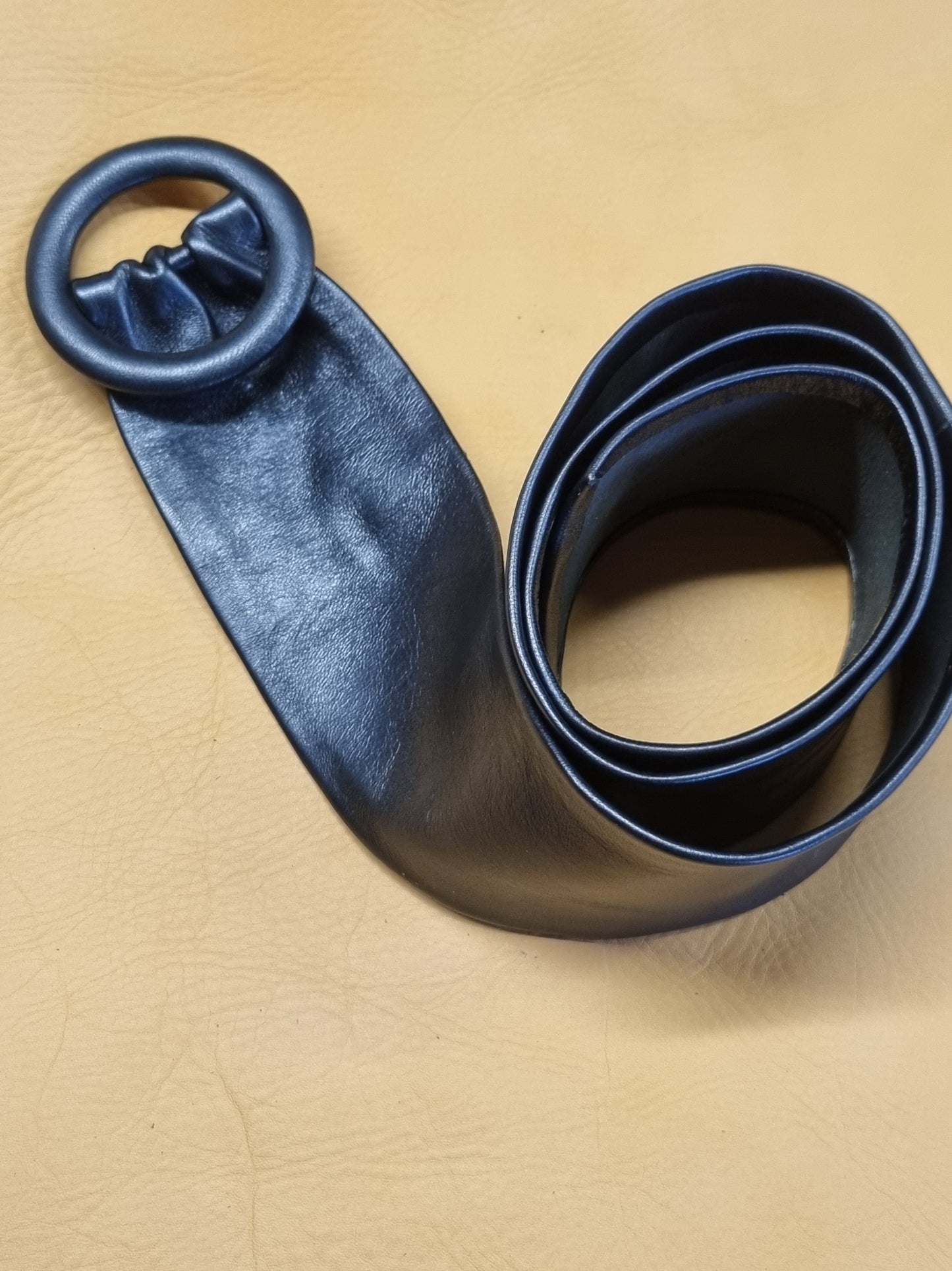 A navy blue leather belt with a wide strap and a large circular leather covered buckle, coiled on a tan textured leather background.