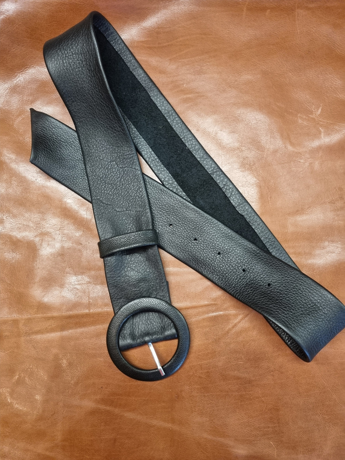 A black leather belt with a large circular ring buckle, laid out on a brown leather background.