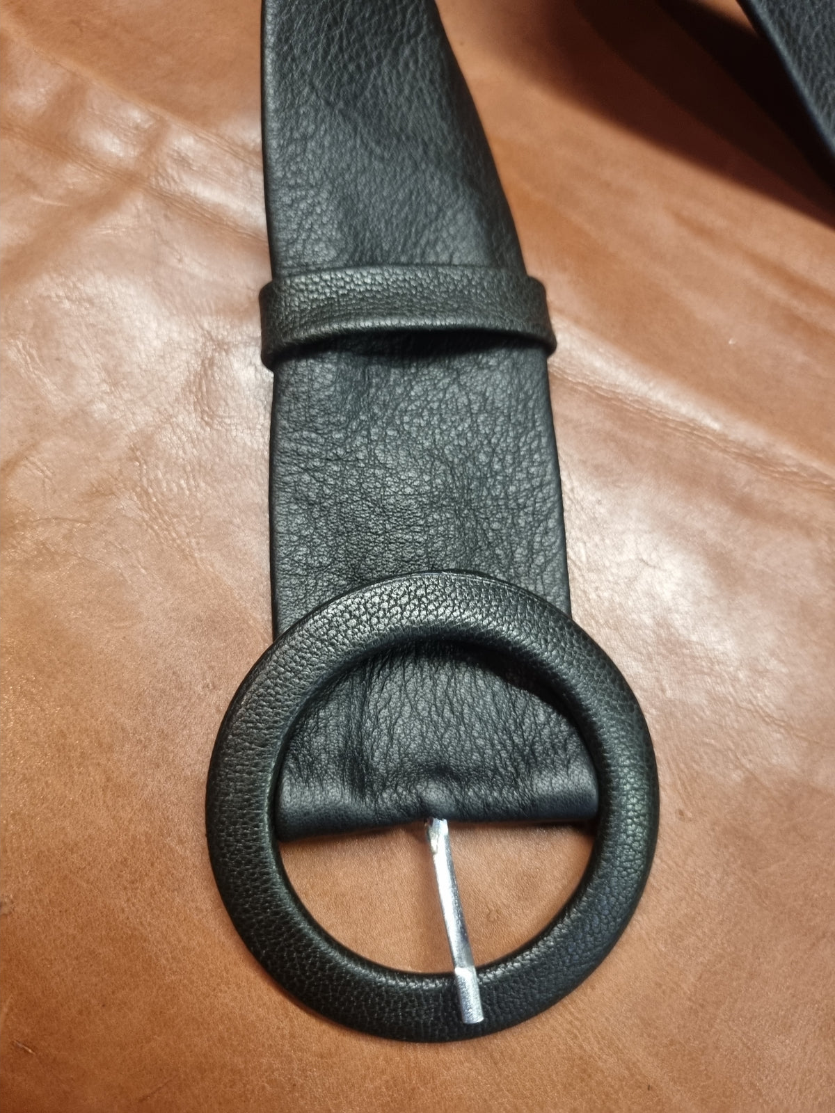 A close-up of a black leather belt with a large circular ring buckle, laid flat on a brown leather surface.