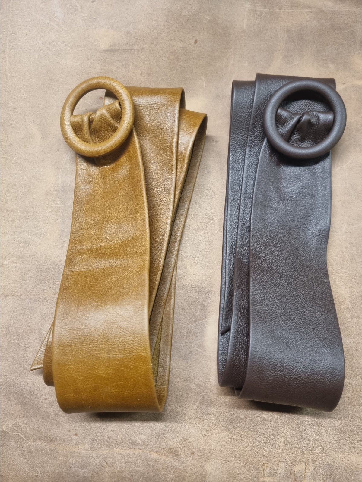 Two rolled leather straps with circular metal ring hardware, one in tan and one in dark charcoal, displayed side by side on a textured neutral surface.