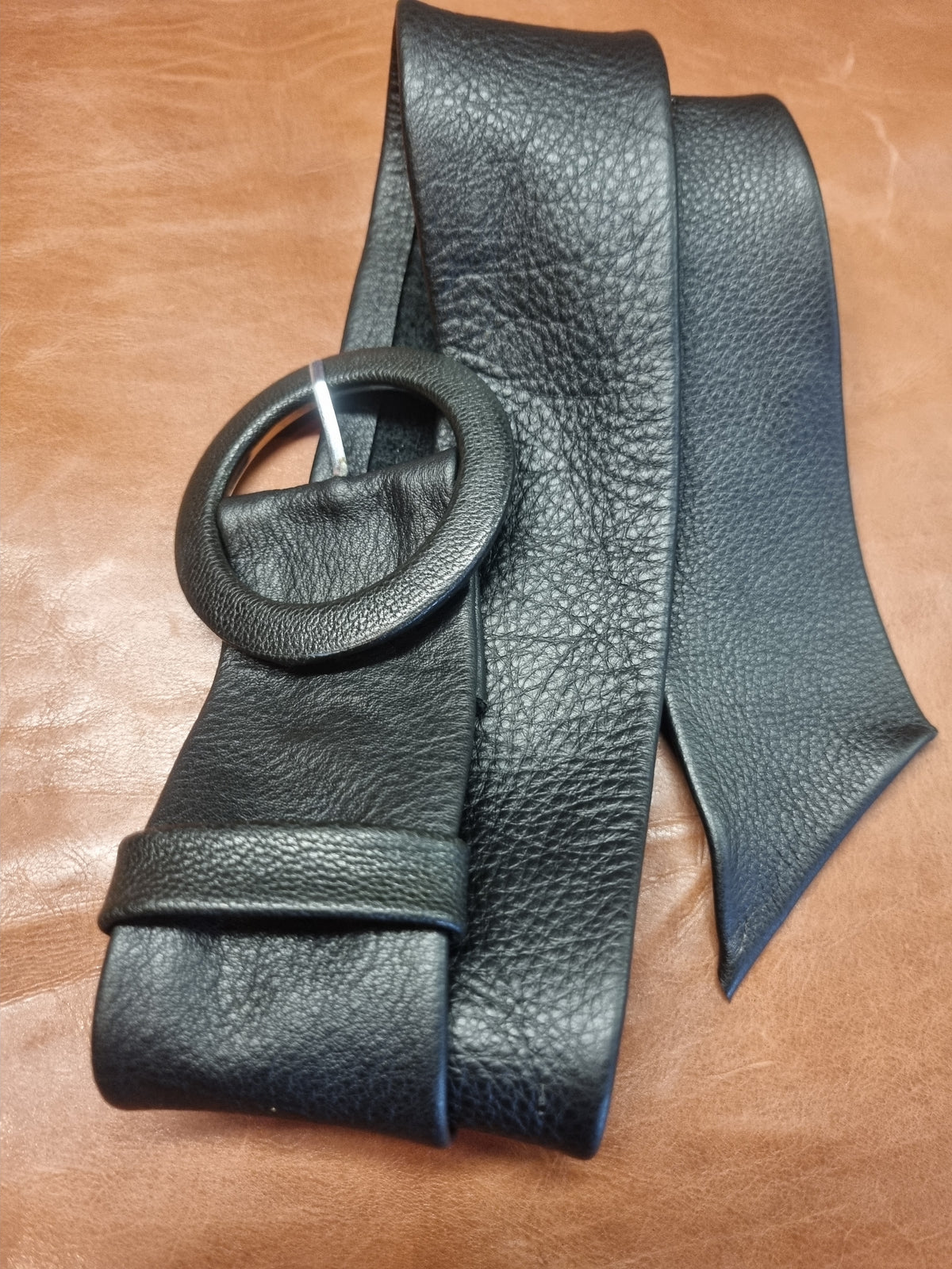 A black pebbled leather belt with a large round metal ring buckle, folded and displayed on a brown leather surface.