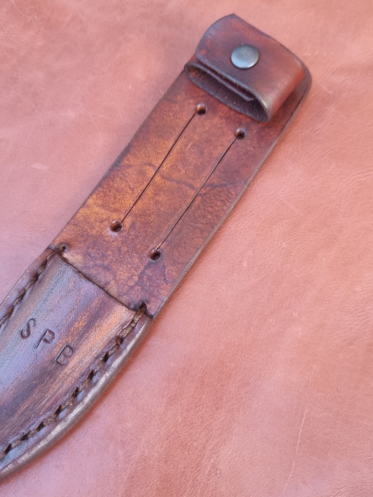 A close-up of a brown leather knife sheath or belt strap laid diagonally on a matching brown leather background. The strap has visible stitching, metal rivets, a snap tab with a metal rivet, and stamped initials 'SP' near the lower end.