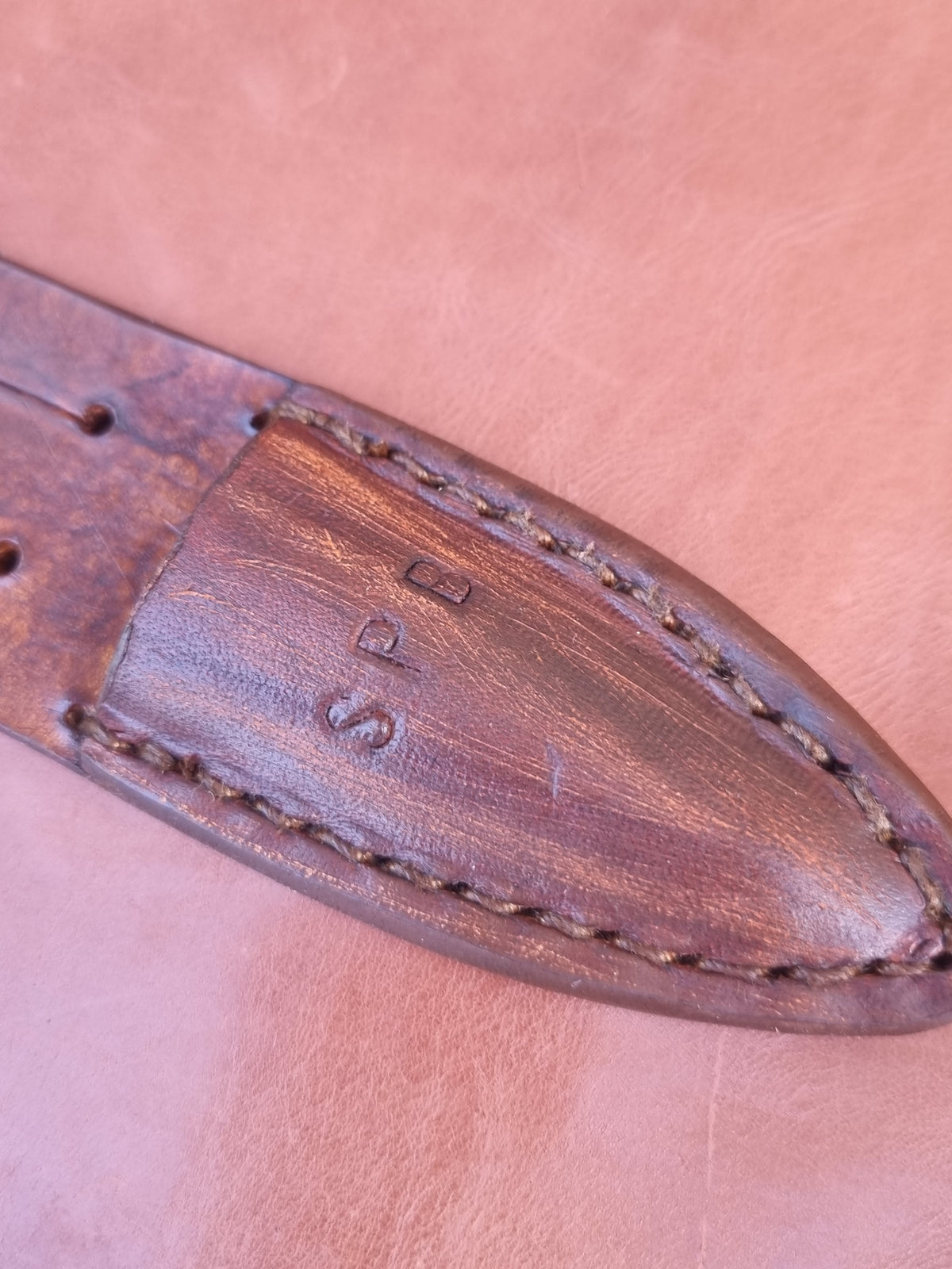 Close-up of a brown leather strap end with visible stitching and stamped initials on a matching tan leather background.