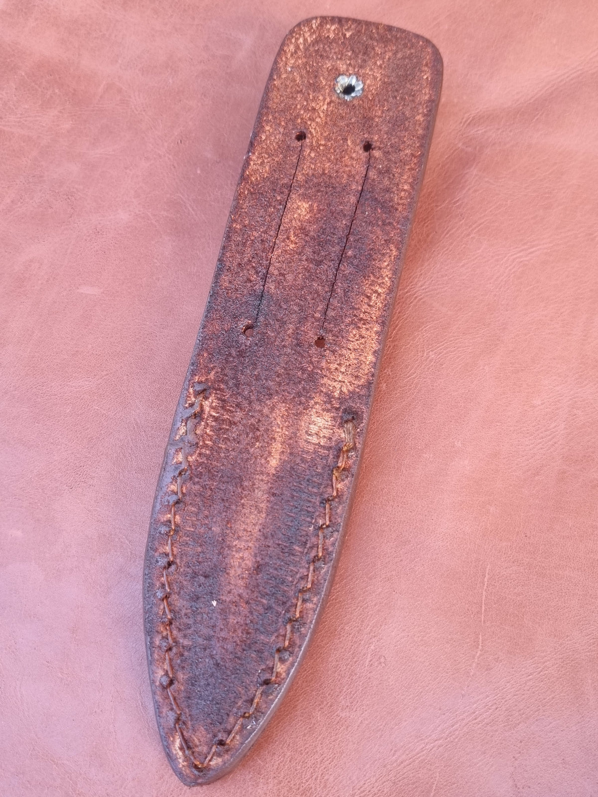 A close-up of a dark brown leather knife sheath with visible stitching and a small metal rivet, placed on a matching brown leather background.