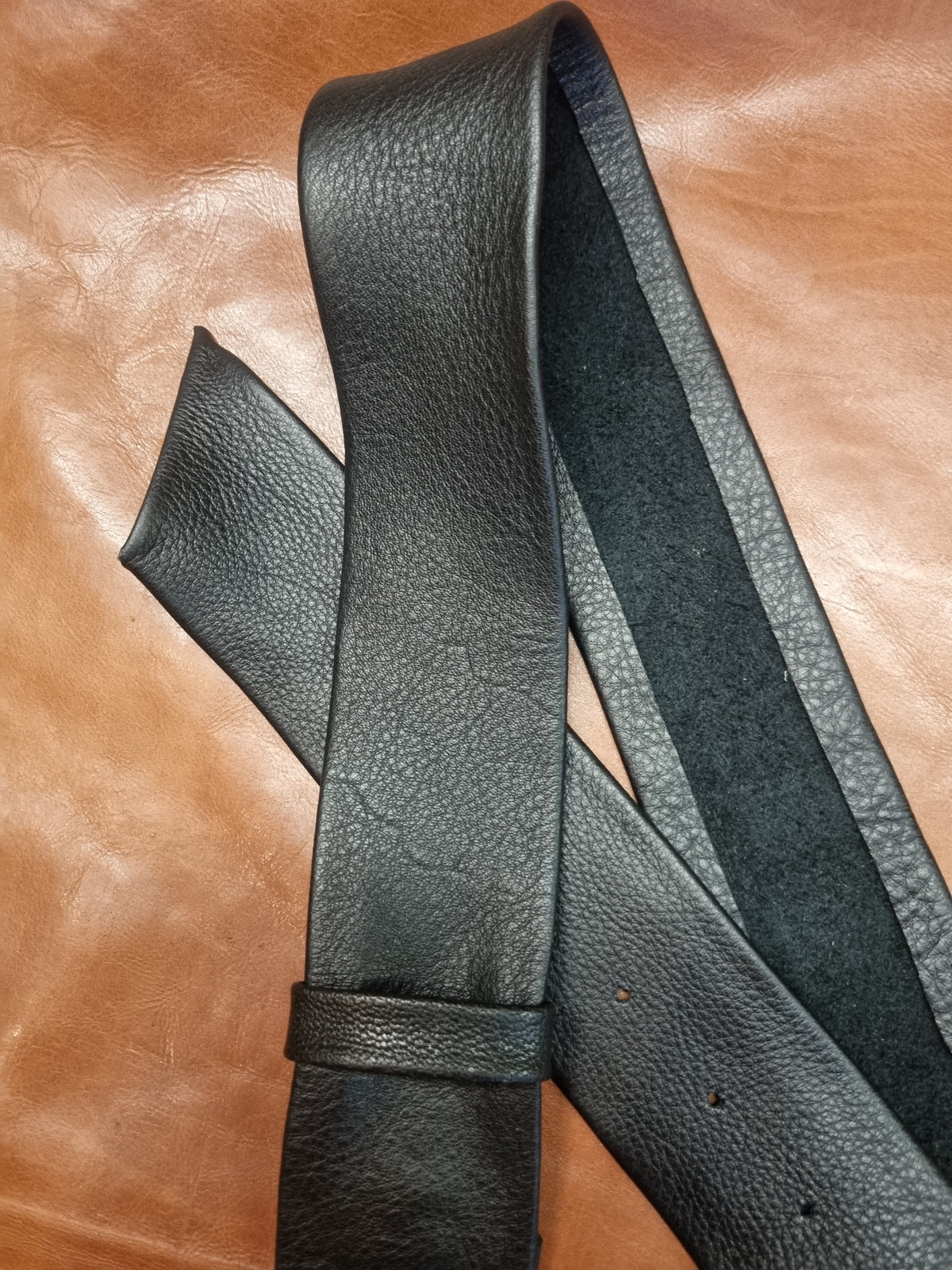 A close-up of a black pebbled leather belt laid diagonally across a brown leather surface, showing both the textured outer strap and the smoother inner lining.