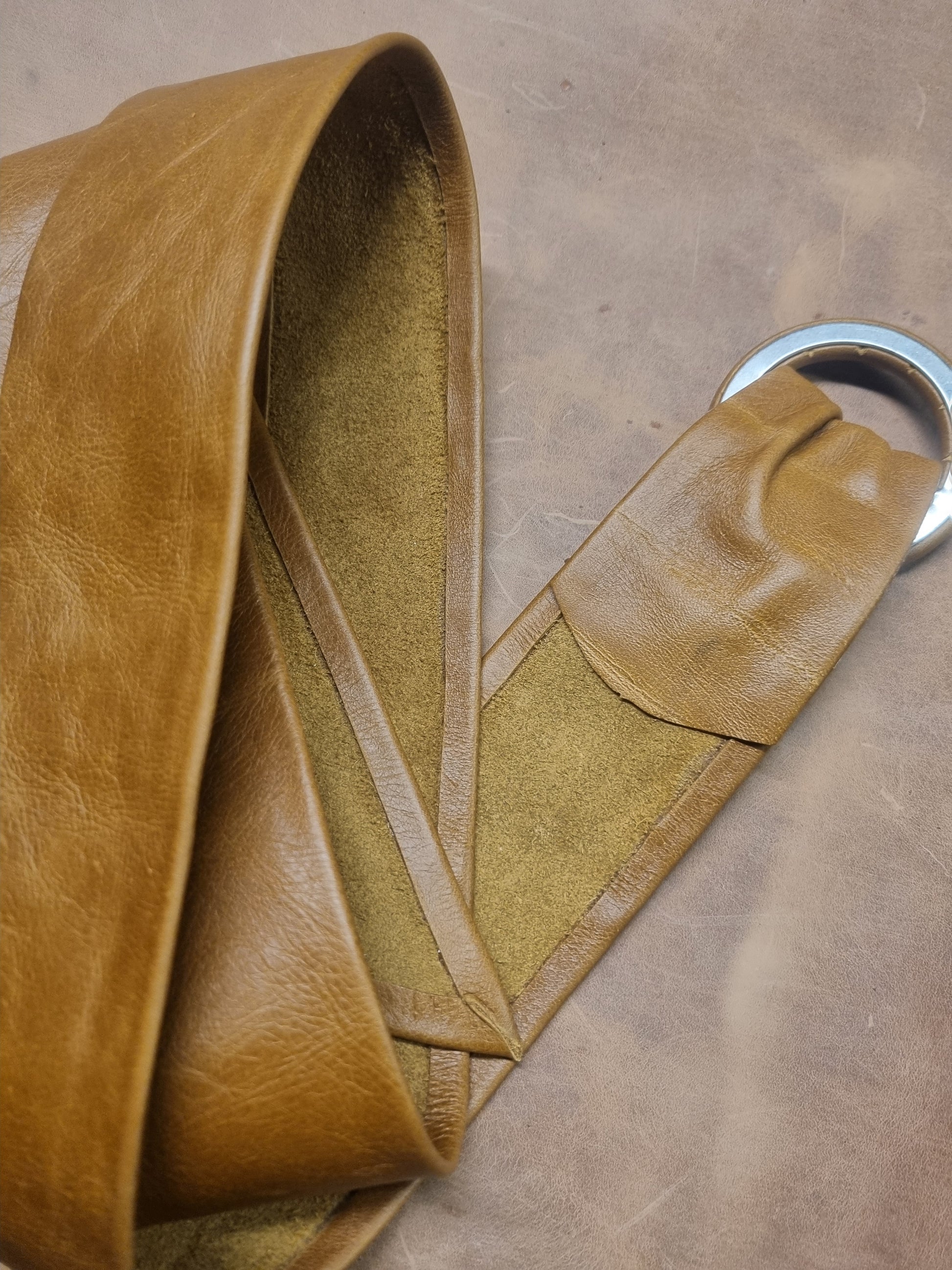 A close-up of a tan leather belt with a silver metal ring, laid on a matching brown textured leather surface.