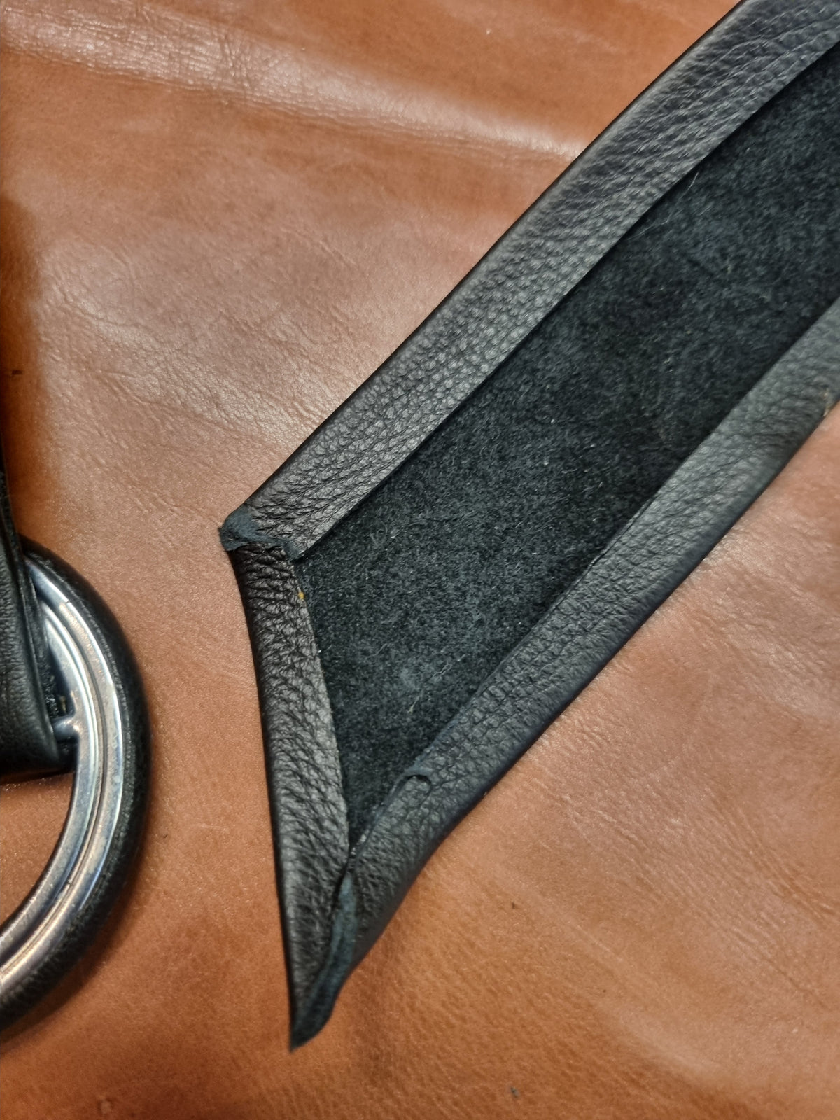 Close-up of a black leather strap with a textured pebbled edge and a dark gray suede or velvety center panel, laid diagonally on a brown leather surface.