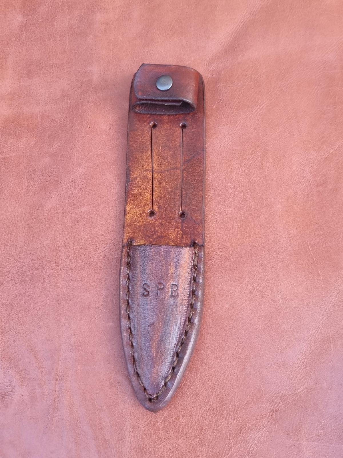 A brown leather knife sheath with a snap closure and stitched edge, featuring the embossed initials 'SPB' on the lower section, displayed on a textured pinkish-brown leather background.