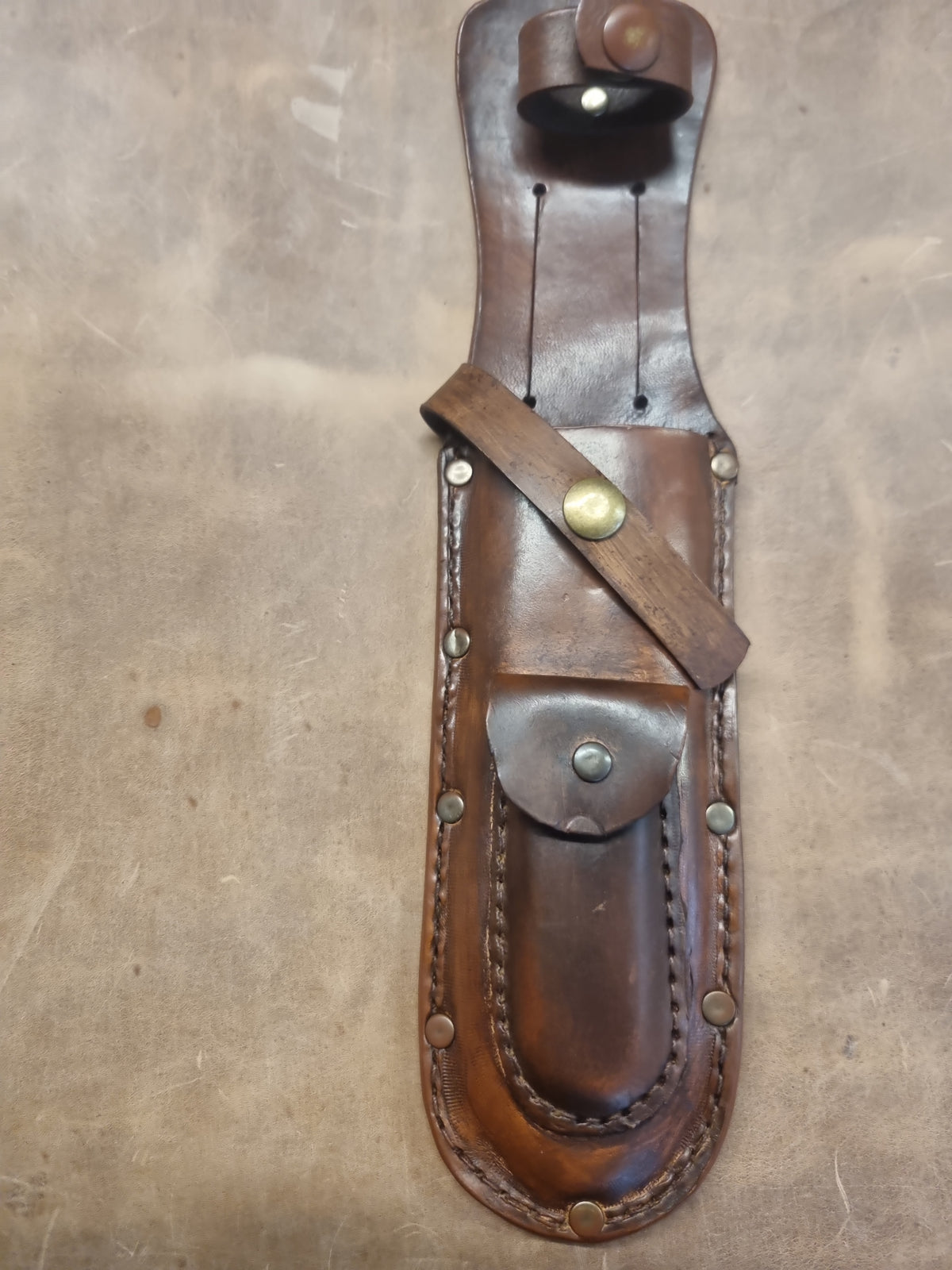 A brown leather knife sheath with brass hardware, laid flat on a textured brown surface.