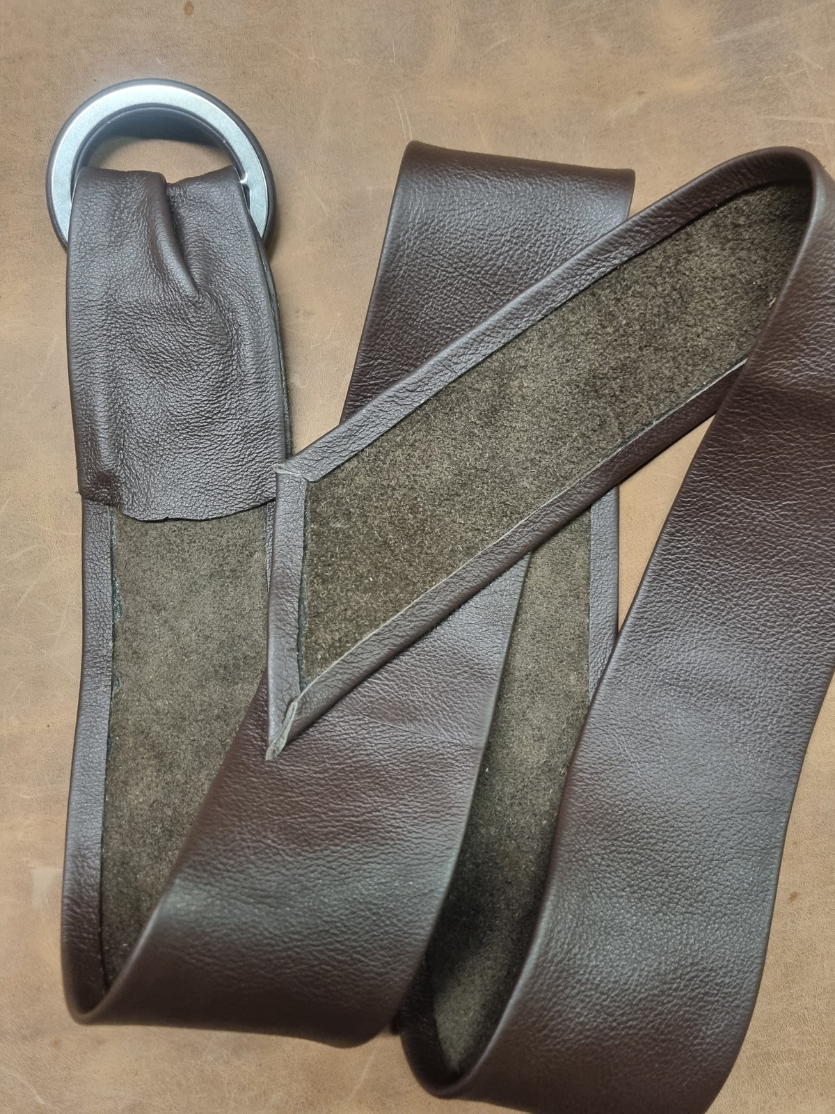 A dark brown leather strap with a silver metal ring and a suede-like brown textured center panel, laid out on a brown surface.