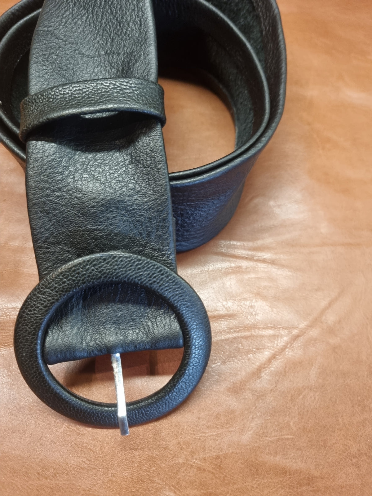 A close-up of a black leather belt with a large round buckle, coiled on a brown leather surface.