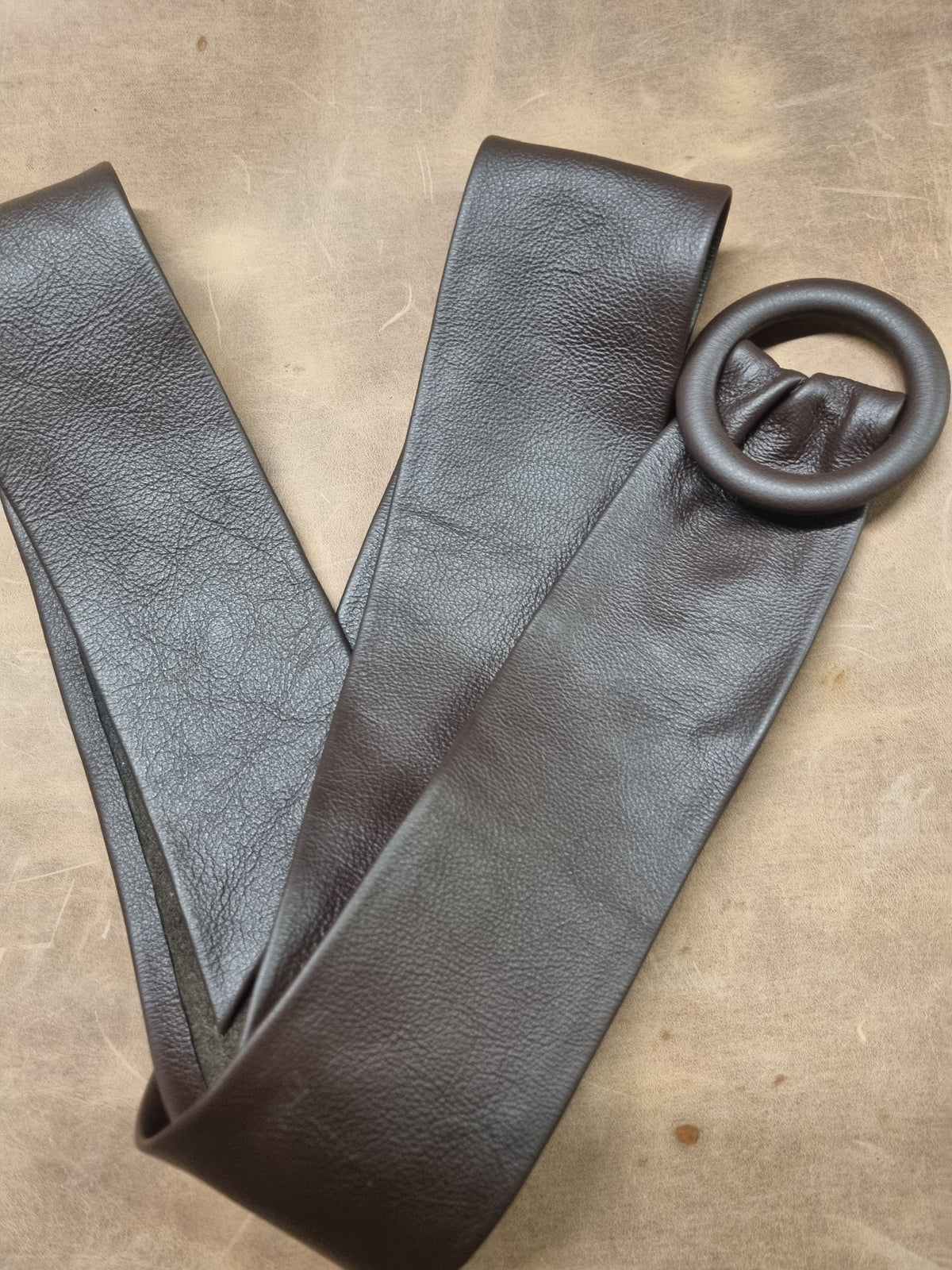 A dark brown textured leather belt with a large matching round metal ring buckle, laid flat on a neutral brown surface.