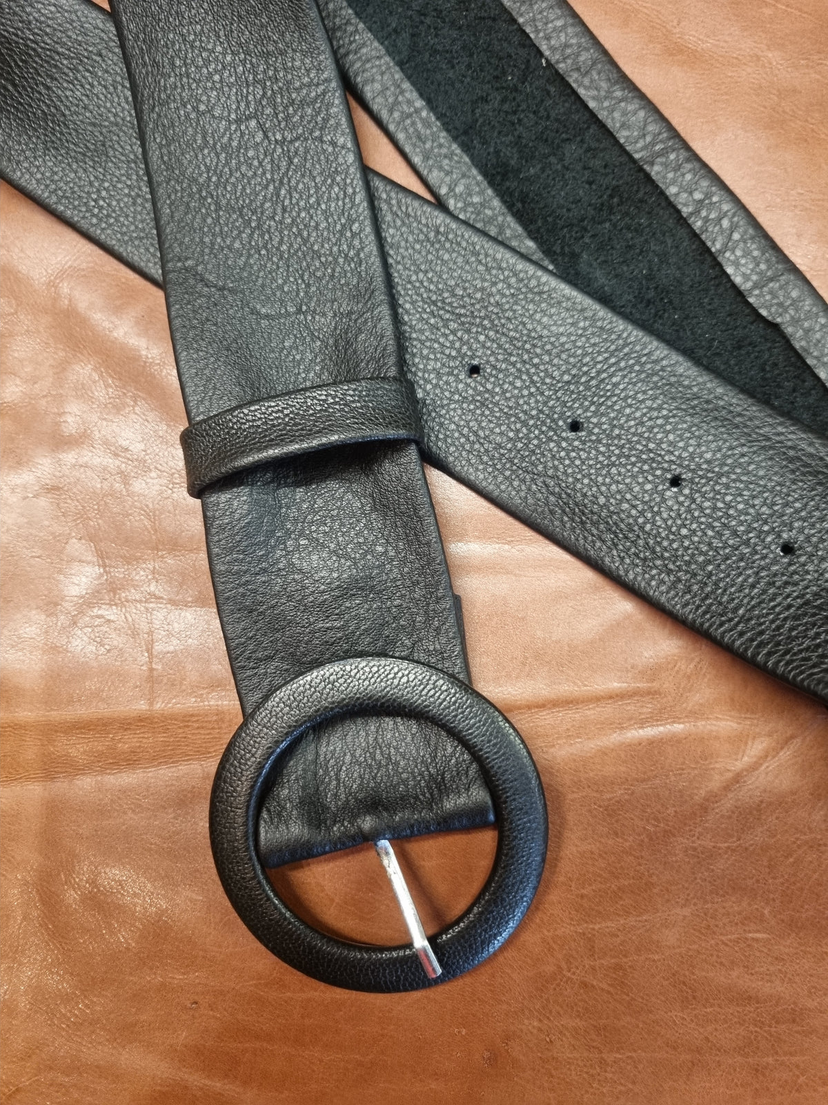 A black leather belt with a large round buckle, laid flat on a brown leather surface.
