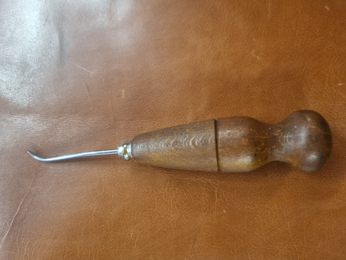 A leatherworking tool with a curved silver metal hook and a wooden handle, resting on a brown leather surface.