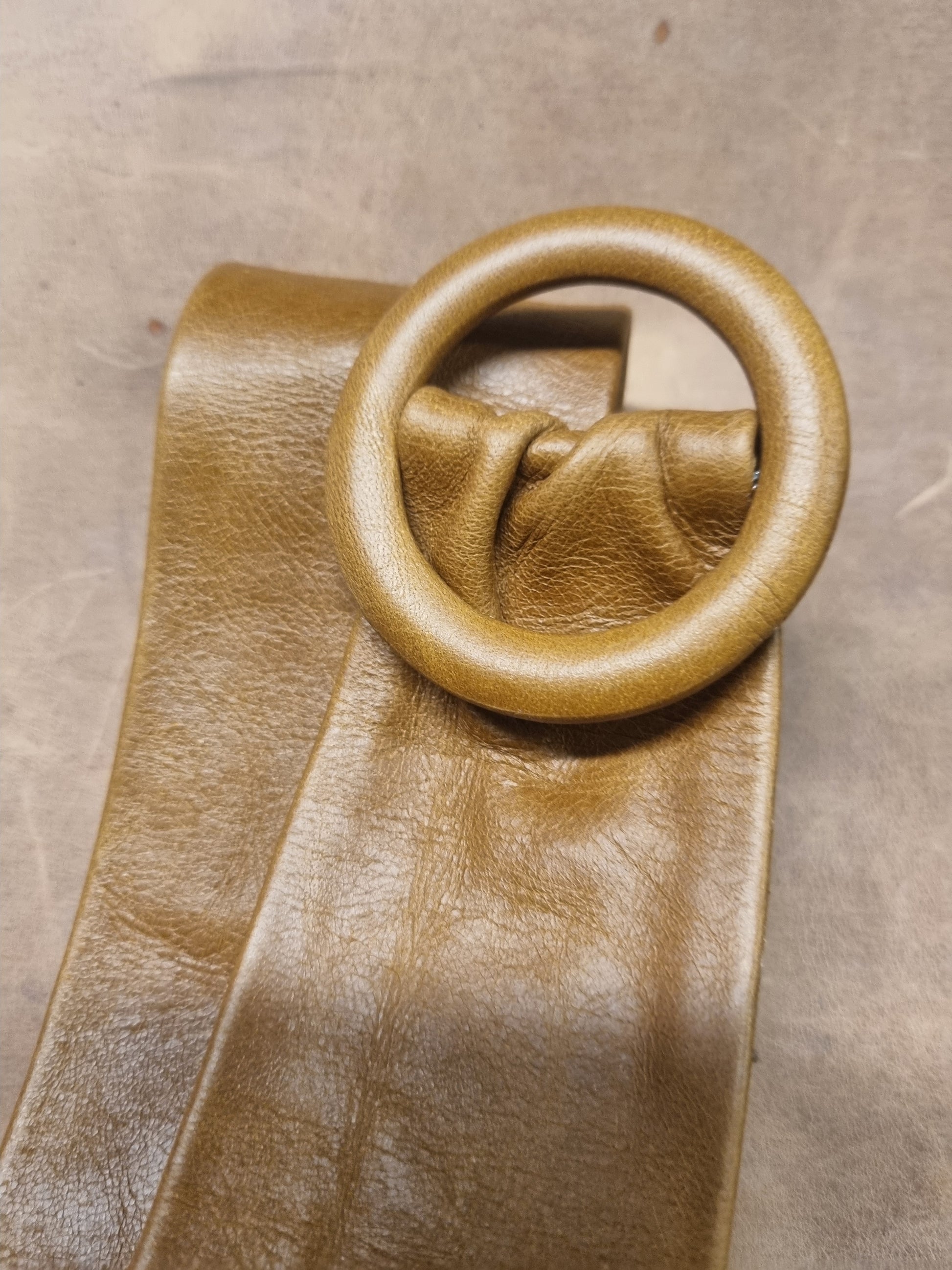 A close-up of a tan leather strap with a large, matching circular ring detail, set against a neutral textured gray background.
