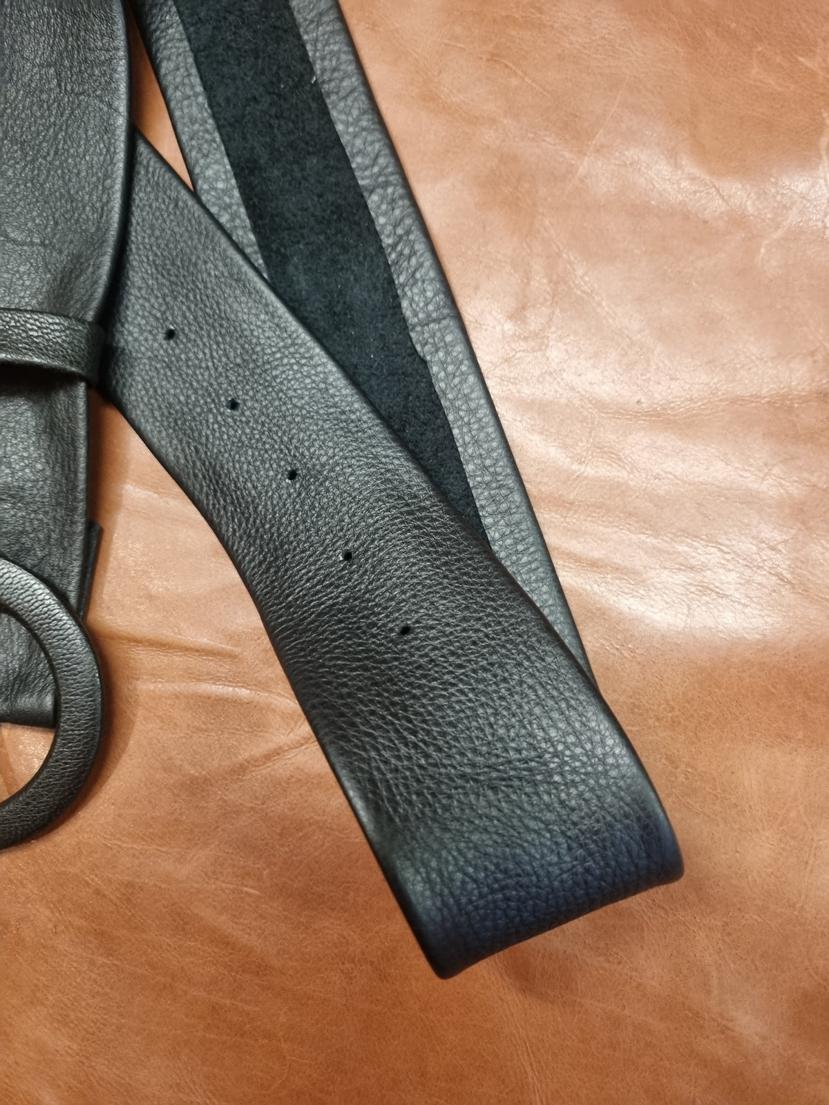 A close-up of a black pebbled leather belt laid diagonally across a brown leather surface, showing the strap texture and belt holes.