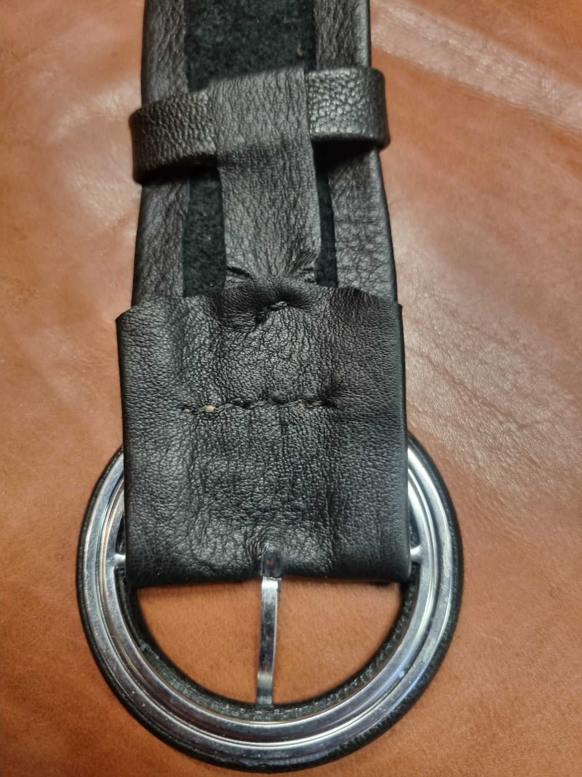 A close-up of a black leather belt with a silver-tone metal buckle, laid flat on a brown leather surface.