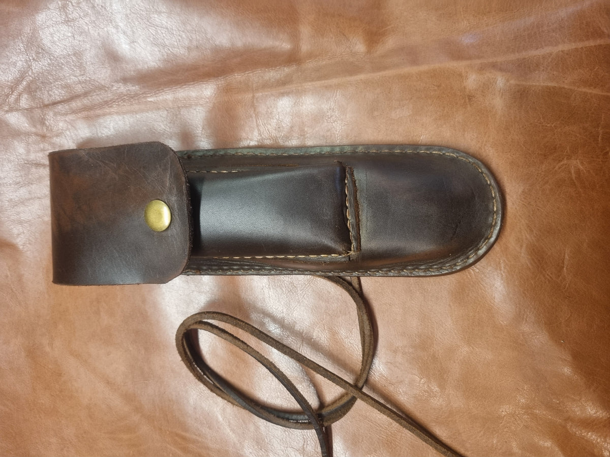 A dark brown leather knife sheath with a brass snap button and a long leather strap, laid flat on a brown leather surface.