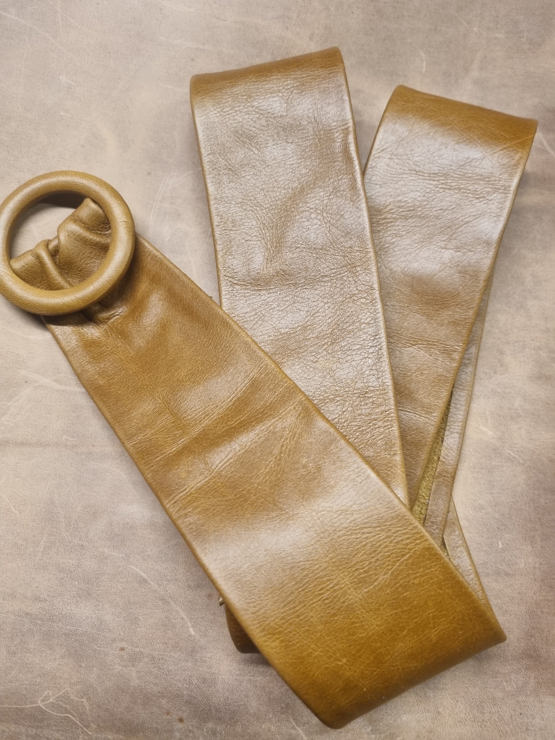 A tan leather belt with a large circular ring buckle, laid flat on a textured gray surface.