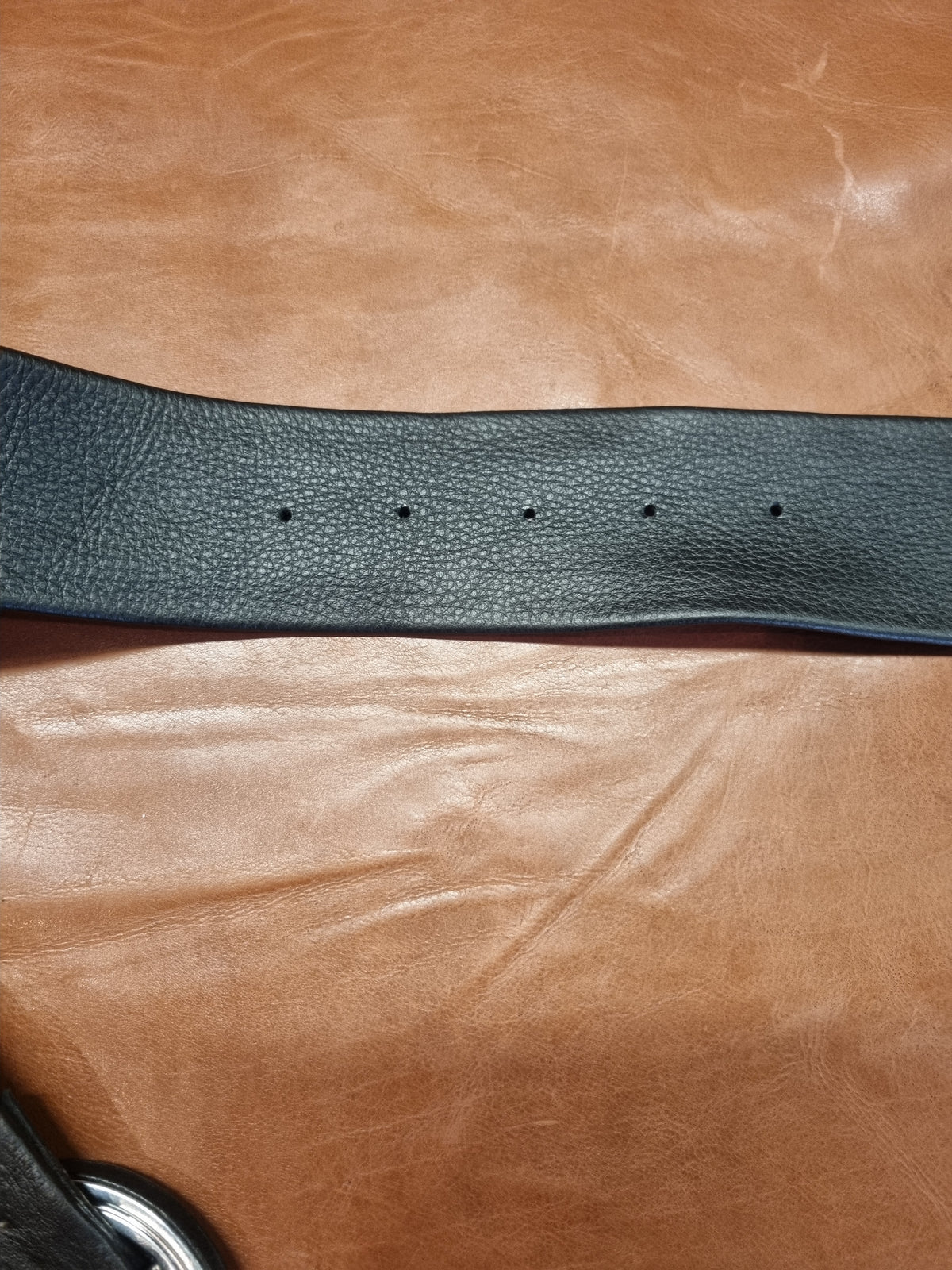 A close-up of a black pebbled leather belt showing buckle holes, laid horizontally across a brown leather background.