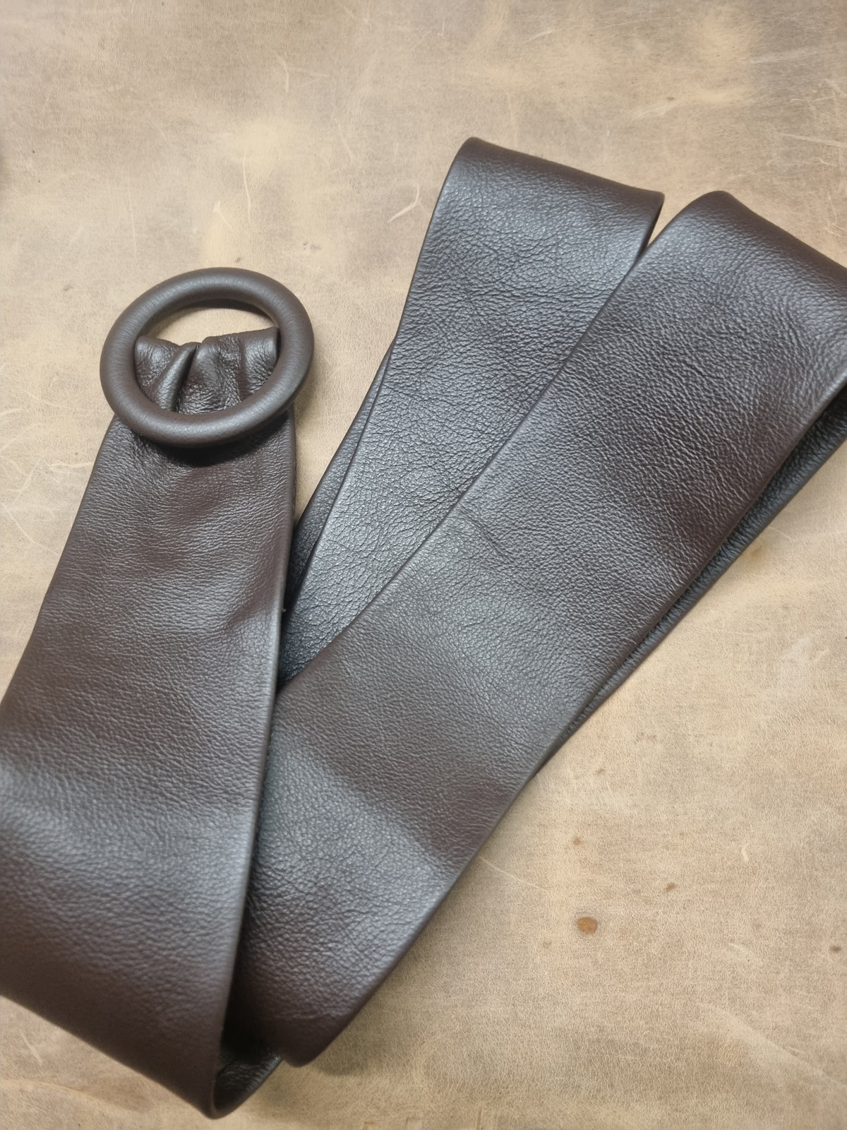 A dark brown textured leather belt with a matching dark brown circular metal ring buckle, laid flat on a textured tan surface.