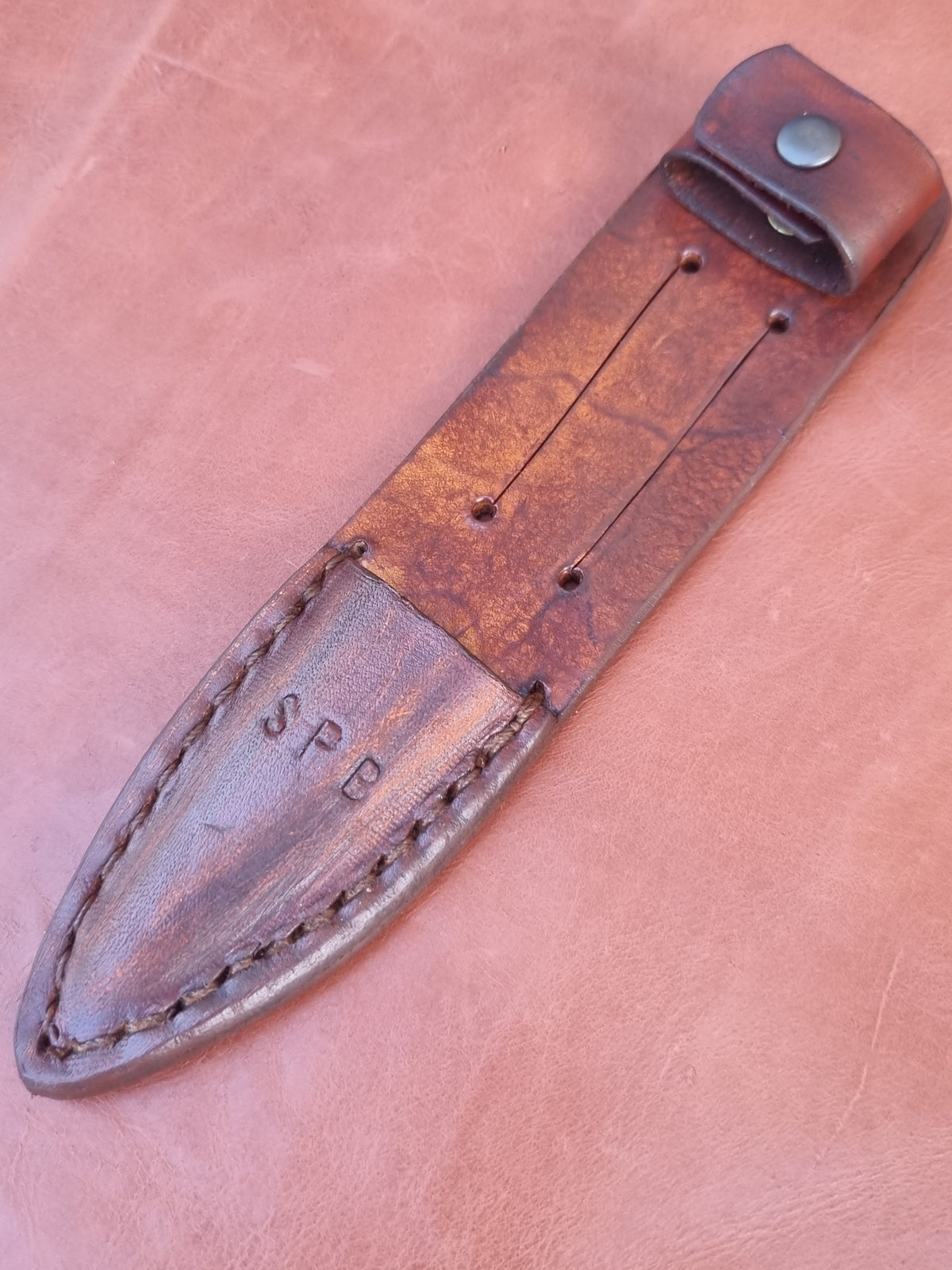 A close-up of a brown leather knife sheath with visible stitching, a snap closure, and the letters 'SPB' stamped on the blade cover, set against a matching leather background.
