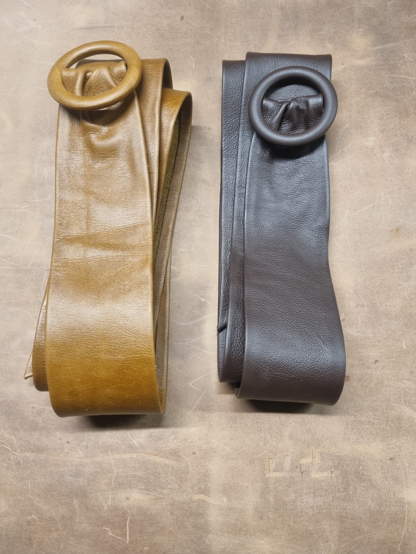 Two leather belts with large circular buckle details, one in tan and one in dark brown, laid flat on a textured brown surface.