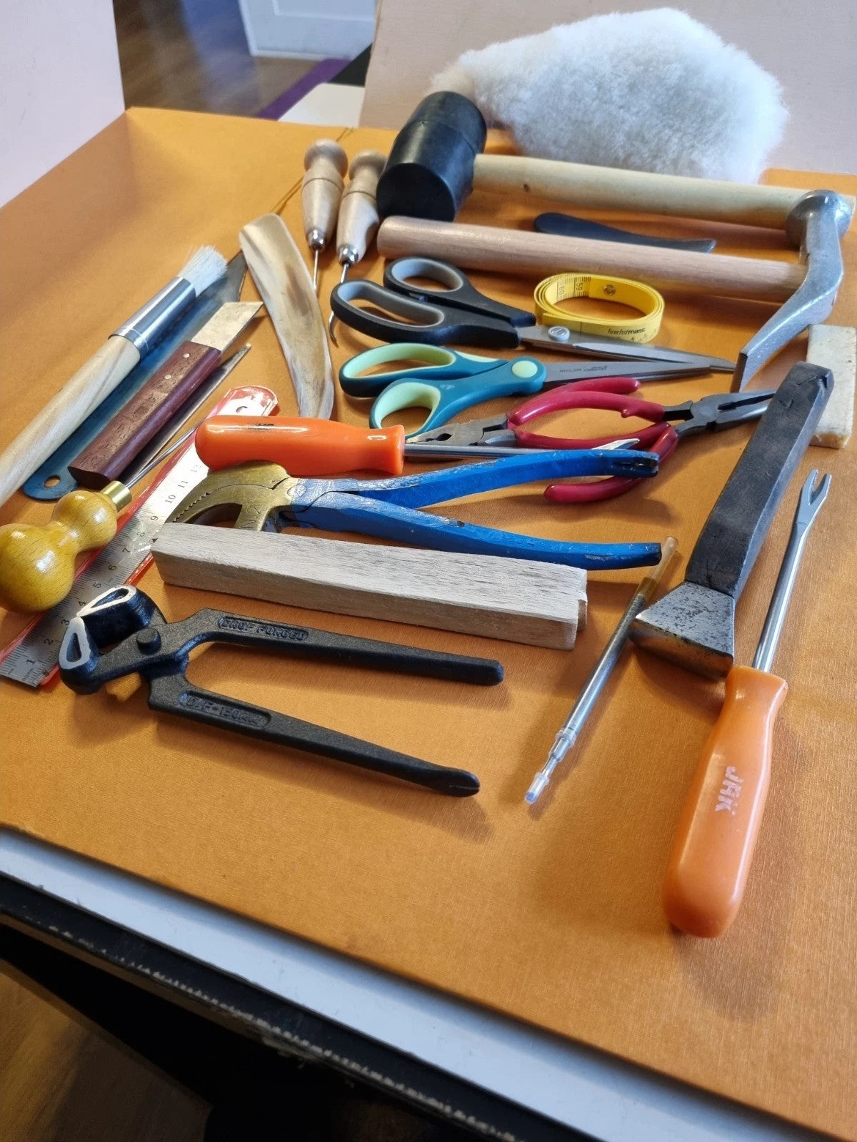 A collection of leatherworking and crafting tools laid out on a tan leather surface, including a hammer, shears, pliers, chisel, rasp, and other hand tools.