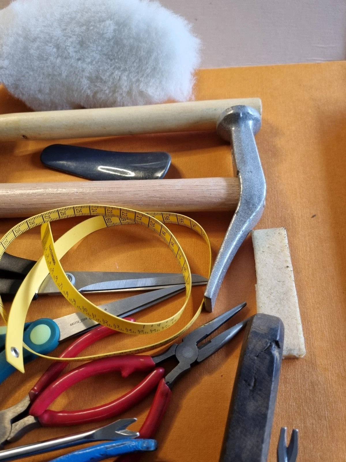 A collection of hand tools and measuring instruments arranged on a wooden surface, including pliers, a tape measure, a hammer, and a white fluffy pom-pom.