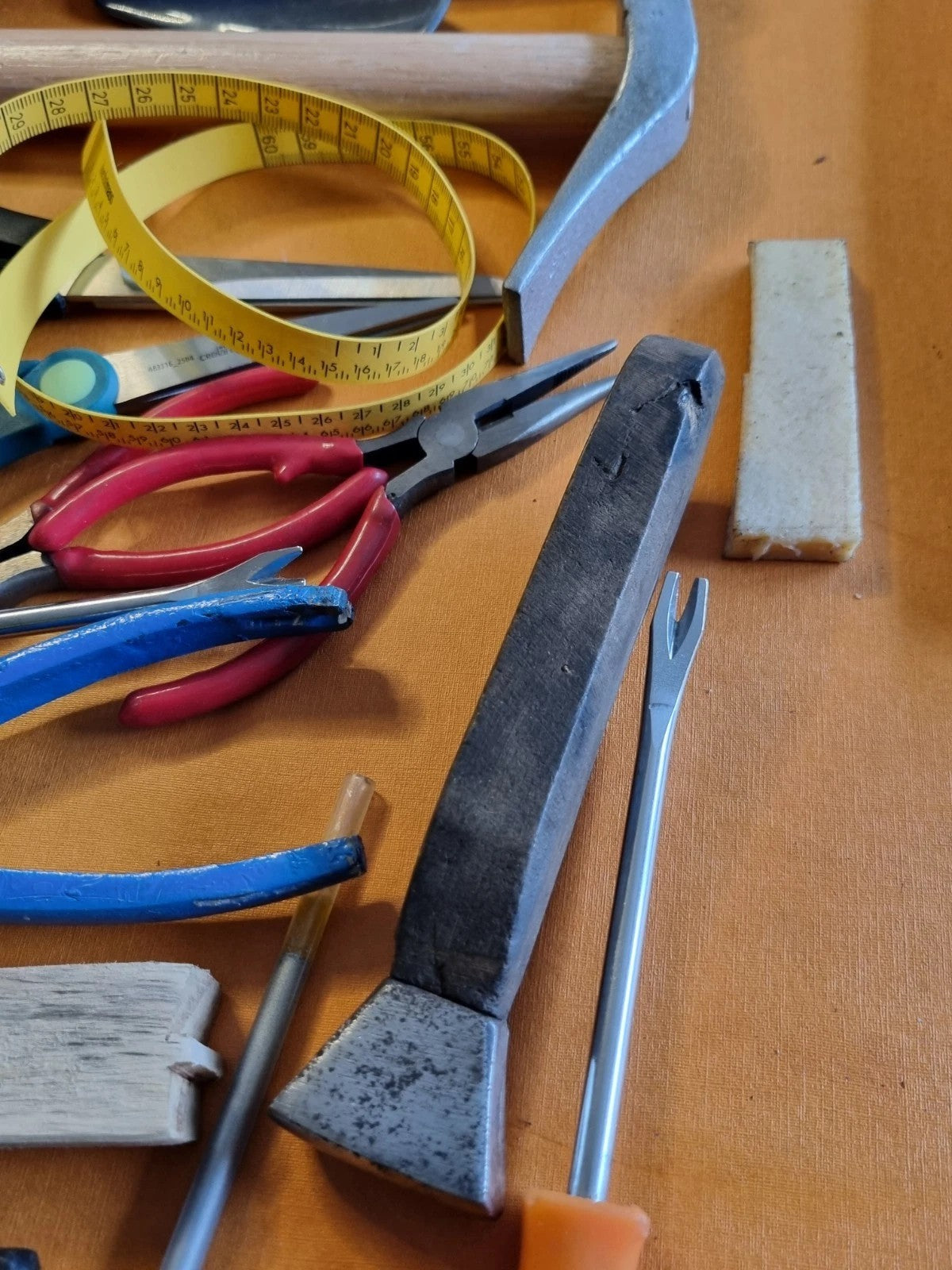 A collection of hand tools including a black-handled axe, pink and blue pliers, a yellow tape measure, and a metal scraper, arranged on a brown wooden surface.