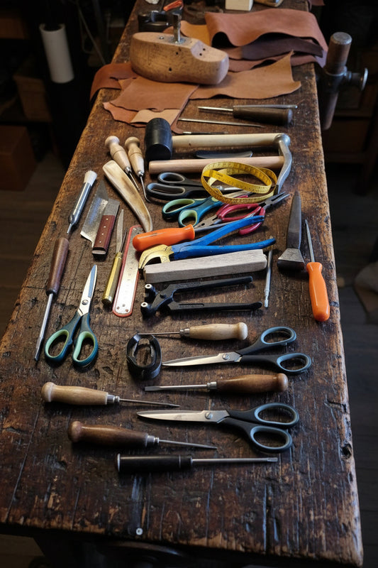 A collection of leatherworking and crafting tools arranged on a tan surface, including a mallet, measuring tape, scissors, pliers, a wooden chisel, a rasp, and other hand tools.